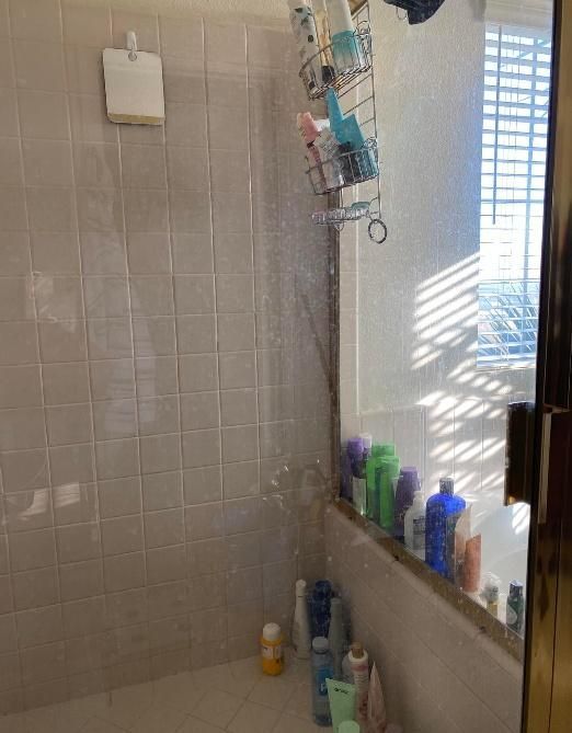 Shower with white tile walls and glass door, filled with toiletries, sunlight visible through window.