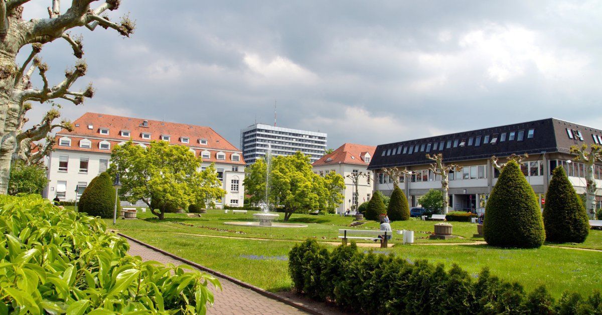 A hospital garden with trimmed hedges, a fountain, trees, benches, and buildings with red and dark roofs in the background.