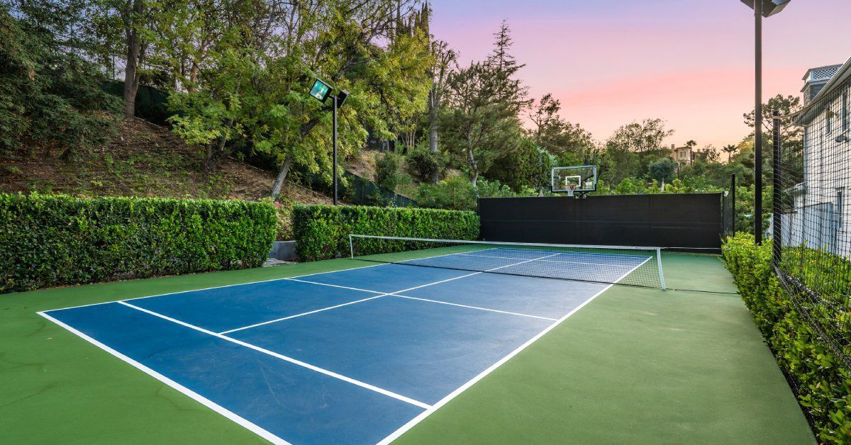 A well-maintained tennis court with blue and green surfaces and white lines, surrounded by lush greenery at sunset.