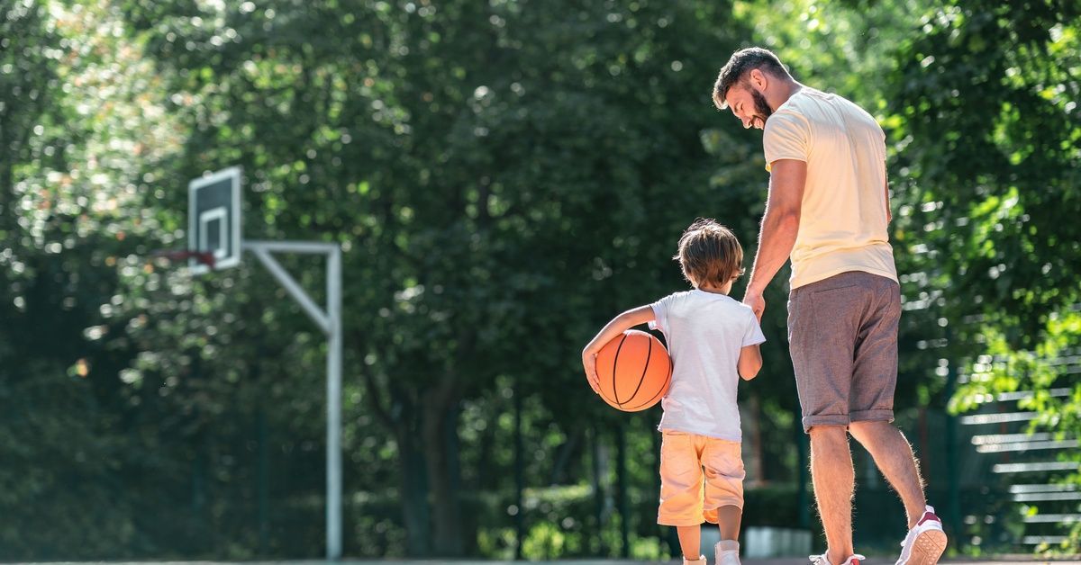 A father walking with his young child on their home sport court. The child is holding a basketball with a hoop up front.
