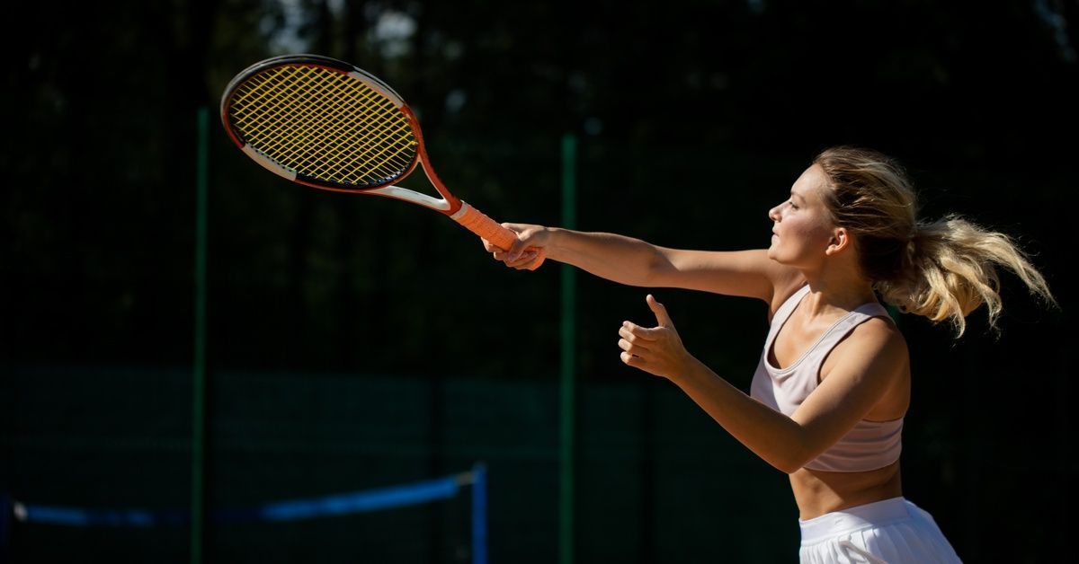 A young tennis player actively swinging her racket at the ball. She's in a ponytail, white tennis skirt, and sport top.