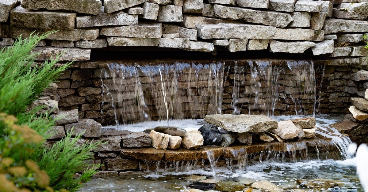 A backyard garden area with a beautiful stone water feature. There's greenery near the water feature as water fills a pond.