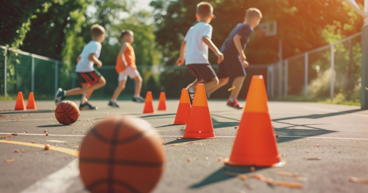 A group of four young boys playing outside on their home athletic court. There are orange cones and two basketballs.