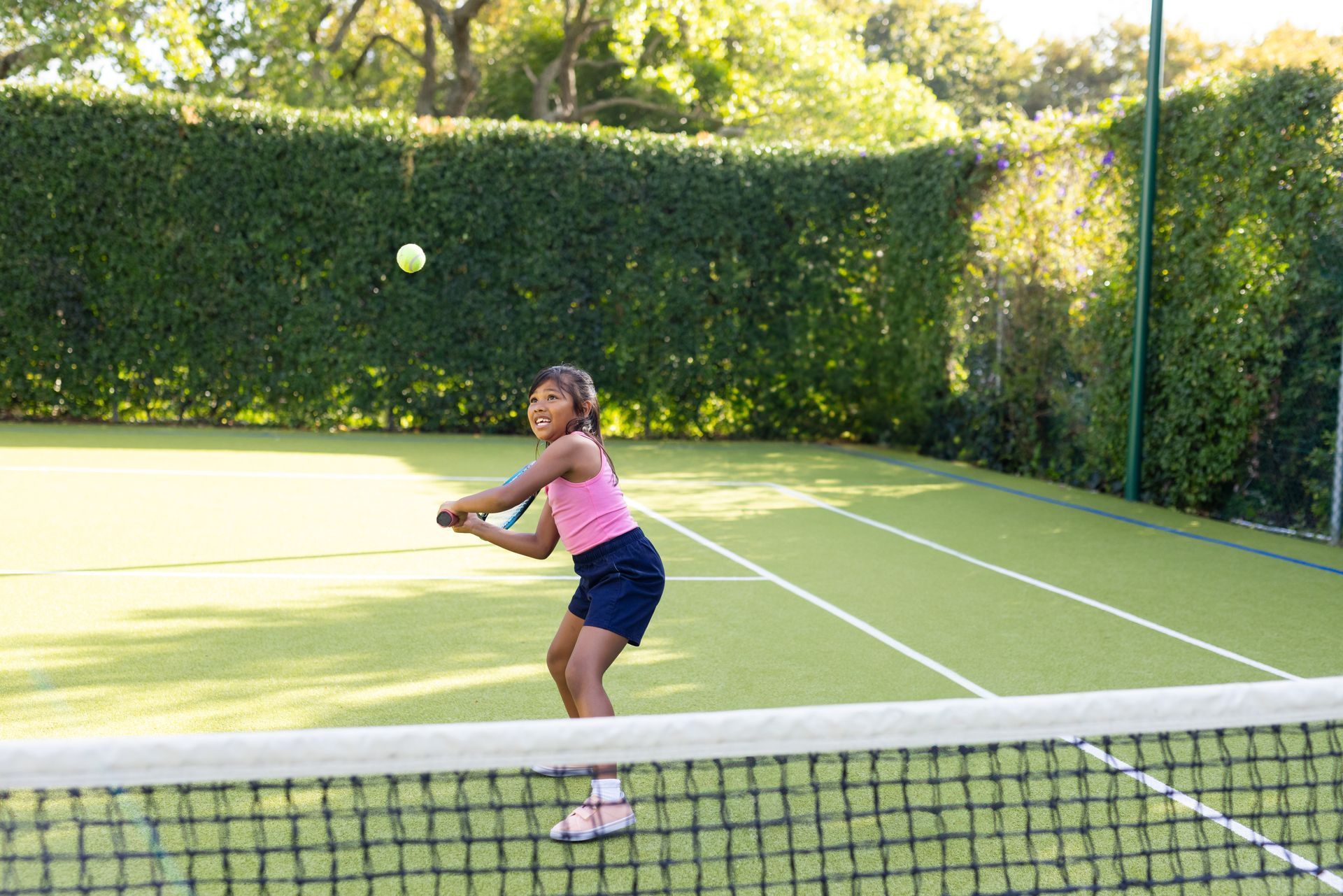 A young girl in a pink tank top playing tennis on her home sports court. The court is surrounded by a tall bush line.