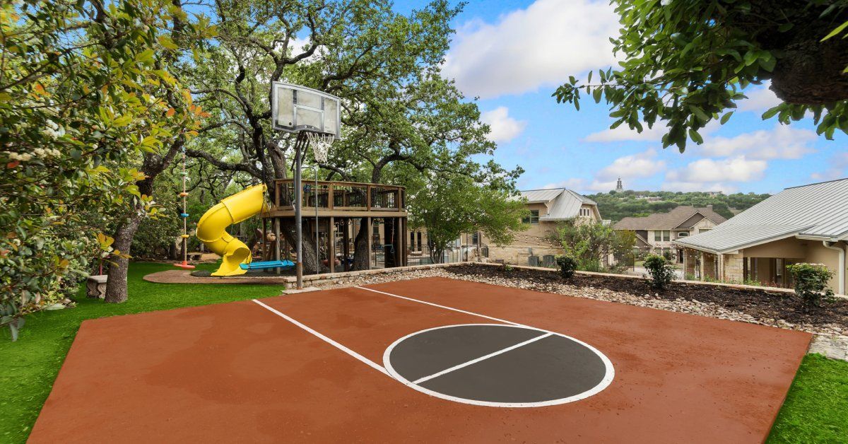 A backyard athletic court with a playground in the back. The court is brown with a basketball hoop.