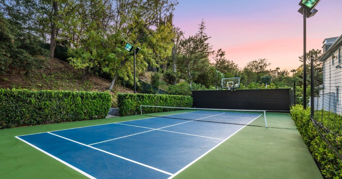 A residential tennis court with blue paint and white lines. Trees surround the court.