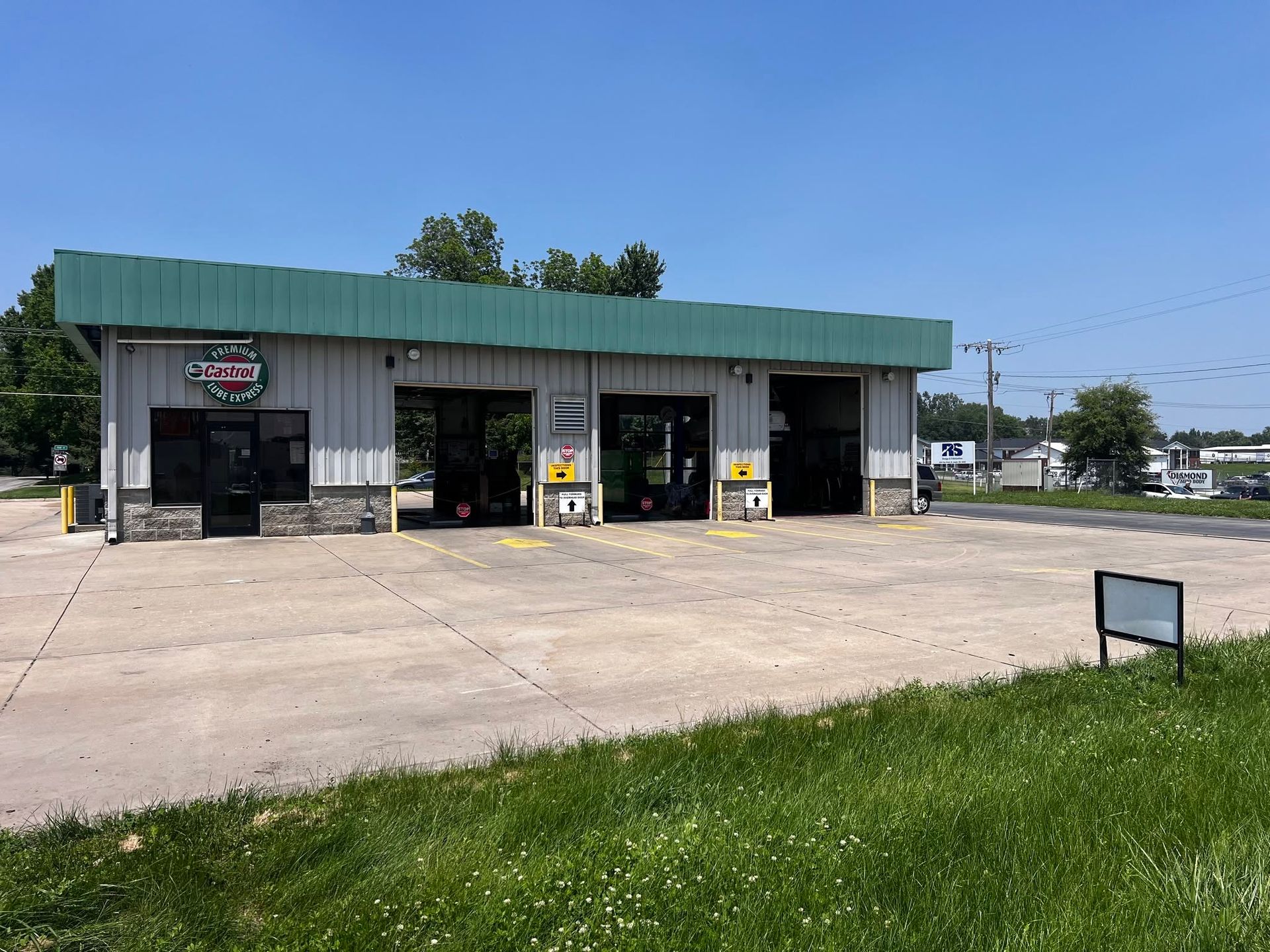 Small roadside storefront with green roof, open garage bays, and a large empty concrete lot. | Castrol Pro-Lube of St. Joe