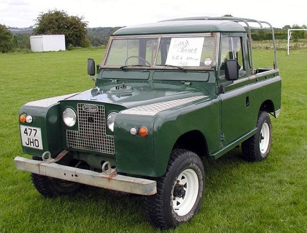 Green vintage Land Rover parked on grass, front-left view with a sign in the windshield.