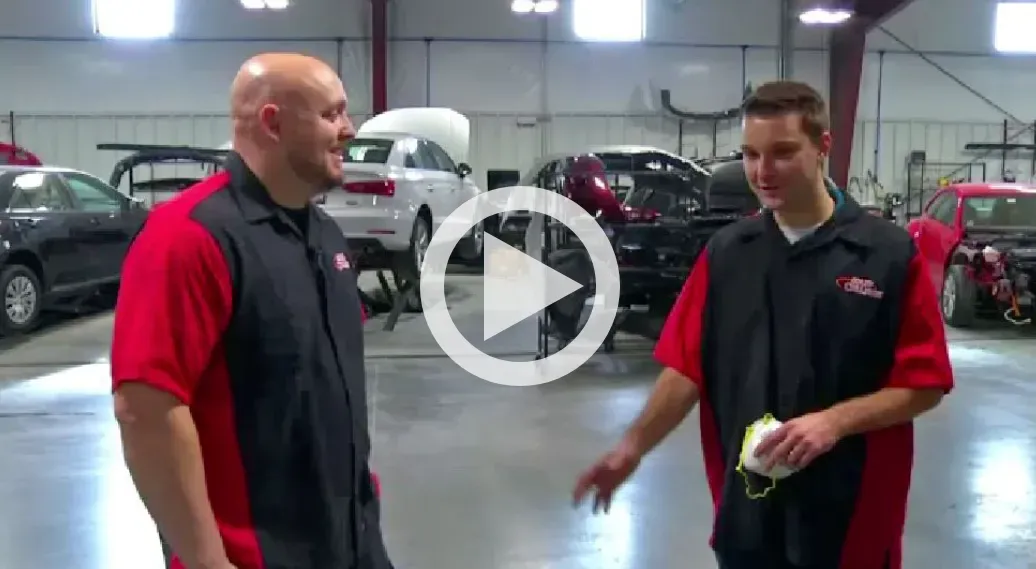Two men in red and black uniforms talking in a car showroom, one holding a tennis ball.