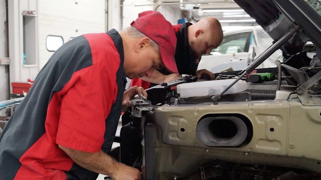 Two mechanics in red and black work under an open car hood in a garage.