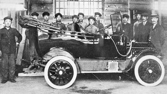 Early automobile with several people standing beside it indoors, black-and-white photo
