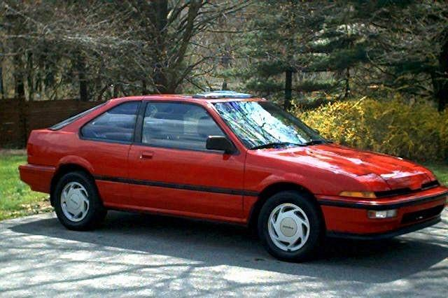 Red compact coupe parked on a driveway beside trees and bushes.
