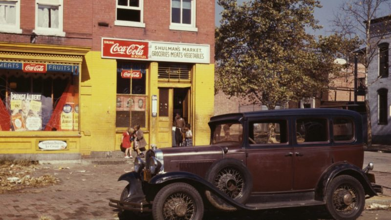Vintage car parked on a street in front of a yellow storefront with a Coca-Cola sign and pedestrians