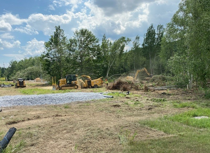 Excavator and construction equipment clearing brush beside a gravel area under a cloudy sky