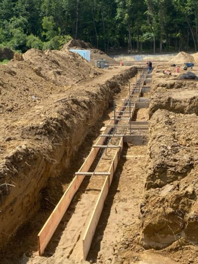 Trenched construction site with wooden forms and a worker in the distance beside a forested area