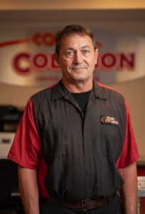 Man standing indoors in a black and red shirt, in front of a blurred company sign.