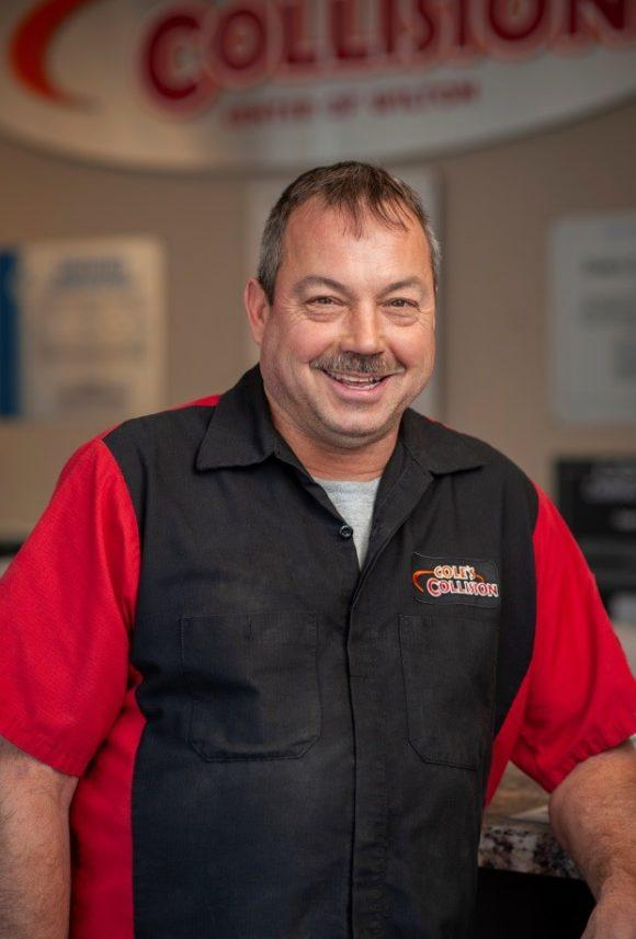 Smiling employee in a red shirt and black vest standing in a collision repair shop
