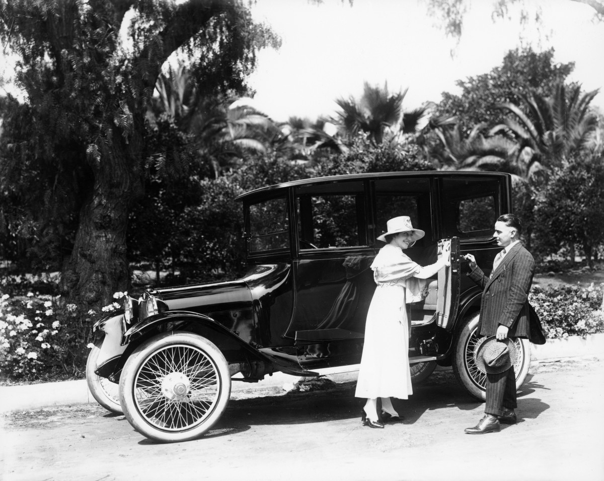 Woman and man beside a vintage car on a palm-lined road, with the woman reaching toward the open door.