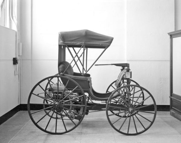 Vintage three-wheeled carriage with a canopy displayed indoors in a museum-like room.