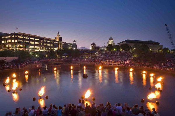 Evening waterfront with floating lanterns, illuminated buildings, and a crowd watching from the shore
