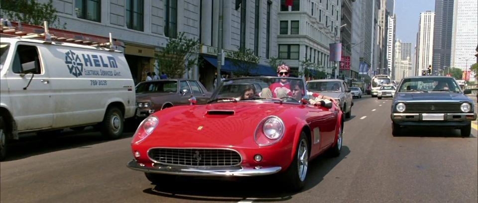 Red convertible sports car driving down a city street with buildings and other cars nearby