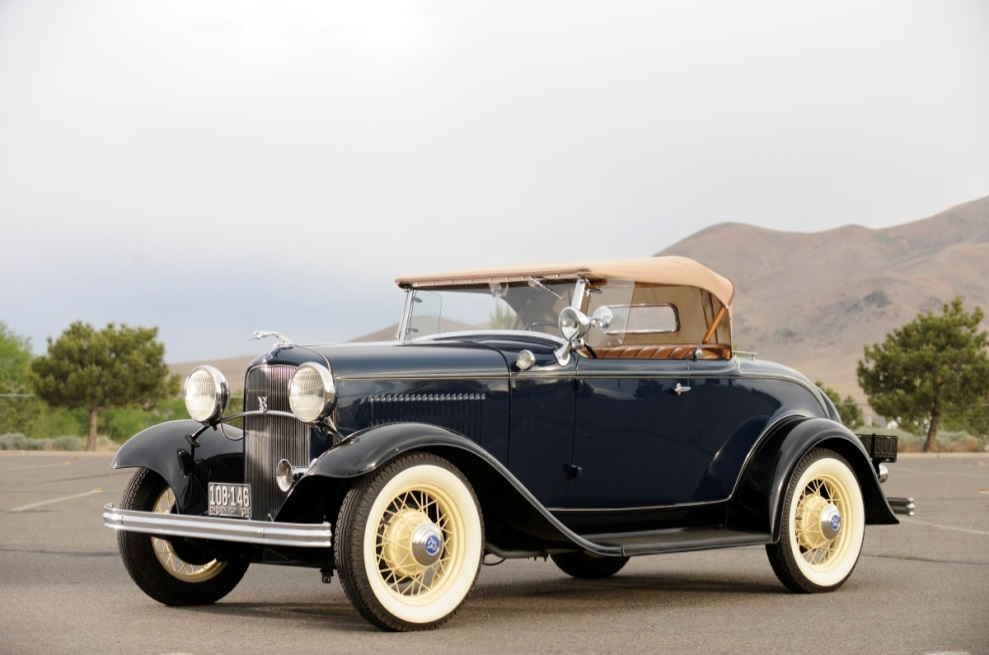 Vintage black convertible car parked outdoors with tan roof and whitewall tires, mountains in the background