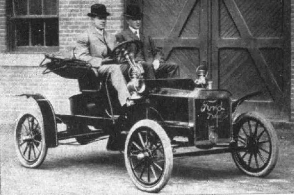 Two people seated in an early open-top automobile in front of a building