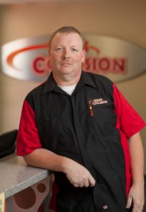 Man standing behind a counter in a red and black shirt, with a blurred “Collision” sign in the background.
