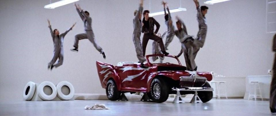 People jumping around a red sports car in a white studio, with tires stacked on the left