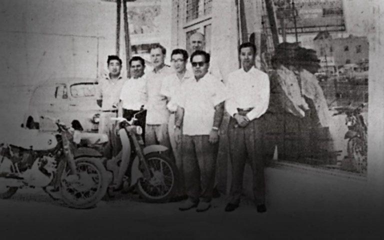 Five men stand beside two motorcycles outside a storefront, one holding an umbrella.