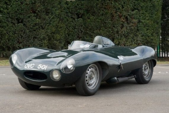 Dark green vintage sports car parked on pavement, front view against a hedge background
