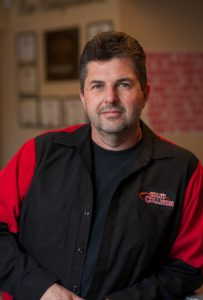 Portrait of a man in a black and red jacket, standing indoors with a slight smile