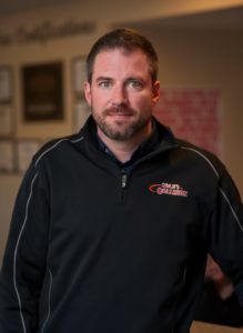 Man in a black zip-up pullover standing indoors against a blurred background.