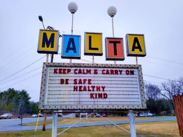 Roadside sign in Malta with colorful letters and a message board reading “Keep calm & carry on. Be safe, healthy, kind.”