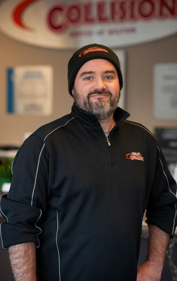 Man in black jacket and beanie standing indoors in front of a Collision auto body sign