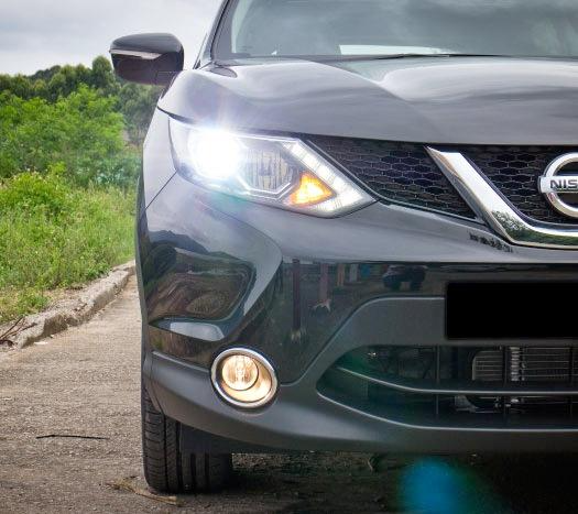 Front view of a dark gray Nissan car with headlights on, parked on a roadside near greenery