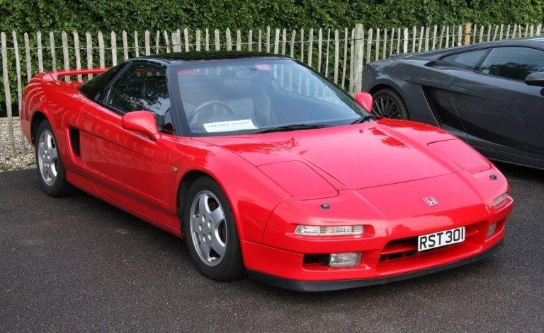 Red sports car parked outdoors beside a black car, with a fence in the background.