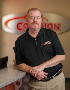 Man in black polo shirt standing by a counter in a lobby with a blurred company sign behind him