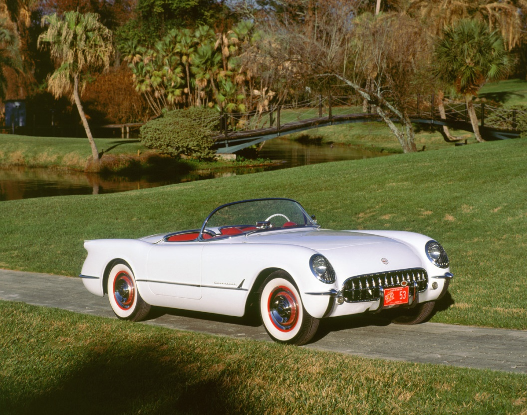 White vintage convertible on a grassy path in a park, with red interior and orange-whitewall tires.