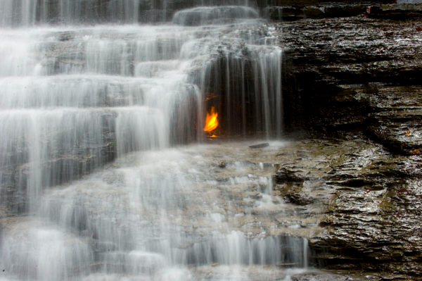 Waterfall cascading over layered rock with a small orange flame burning at center.