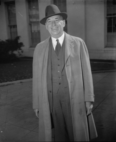 Smiling man in a hat and long coat standing outside a building.