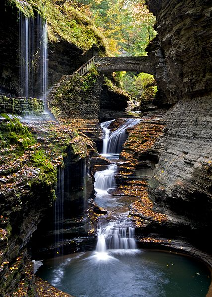 Waterfall cascading through a rocky gorge into a deep pool, with a stone bridge above and autumn foliage.