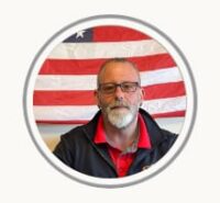 Portrait of a man with glasses and a gray beard in front of a U.S. flag