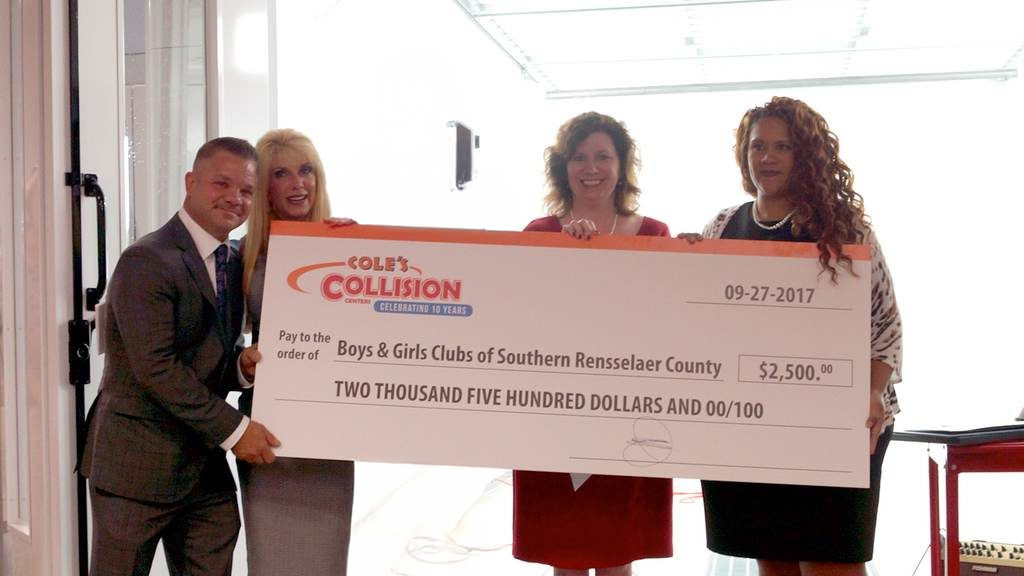 Four people hold a large ceremonial check in a bright hallway.