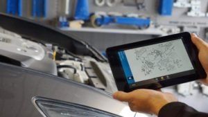 Hand holding a tablet showing a car diagnostic screen under the open hood of a vehicle