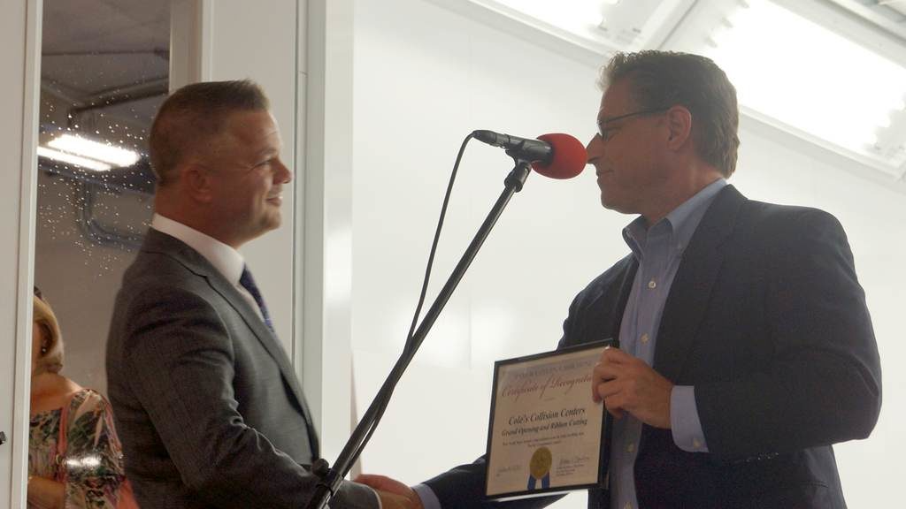Two men in suits shake hands during an award ceremony, one holding a framed certificate at a microphone.