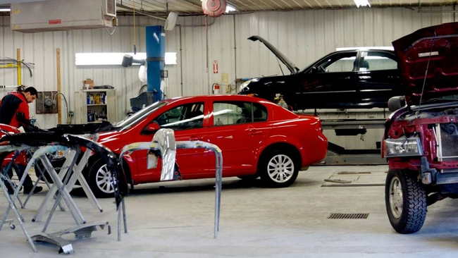 Red sedan in auto repair shop with hood open and mechanic working nearby; lifted black car in background