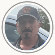 Man wearing a dark cap and jacket, standing beside a silver car outdoors.
