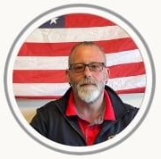 Man with glasses and gray beard in front of a waving American flag background.