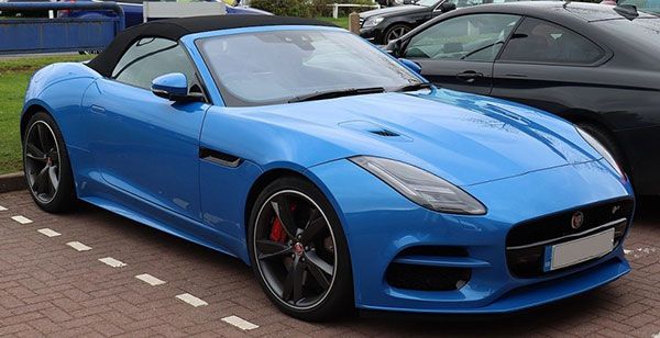 Blue Jaguar sports car parked on a brick street beside a black car.